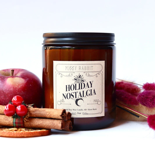 Candle labeled 'Holiday Nostalgia' with apples, cinnamon sticks, and dried flowers on a white background