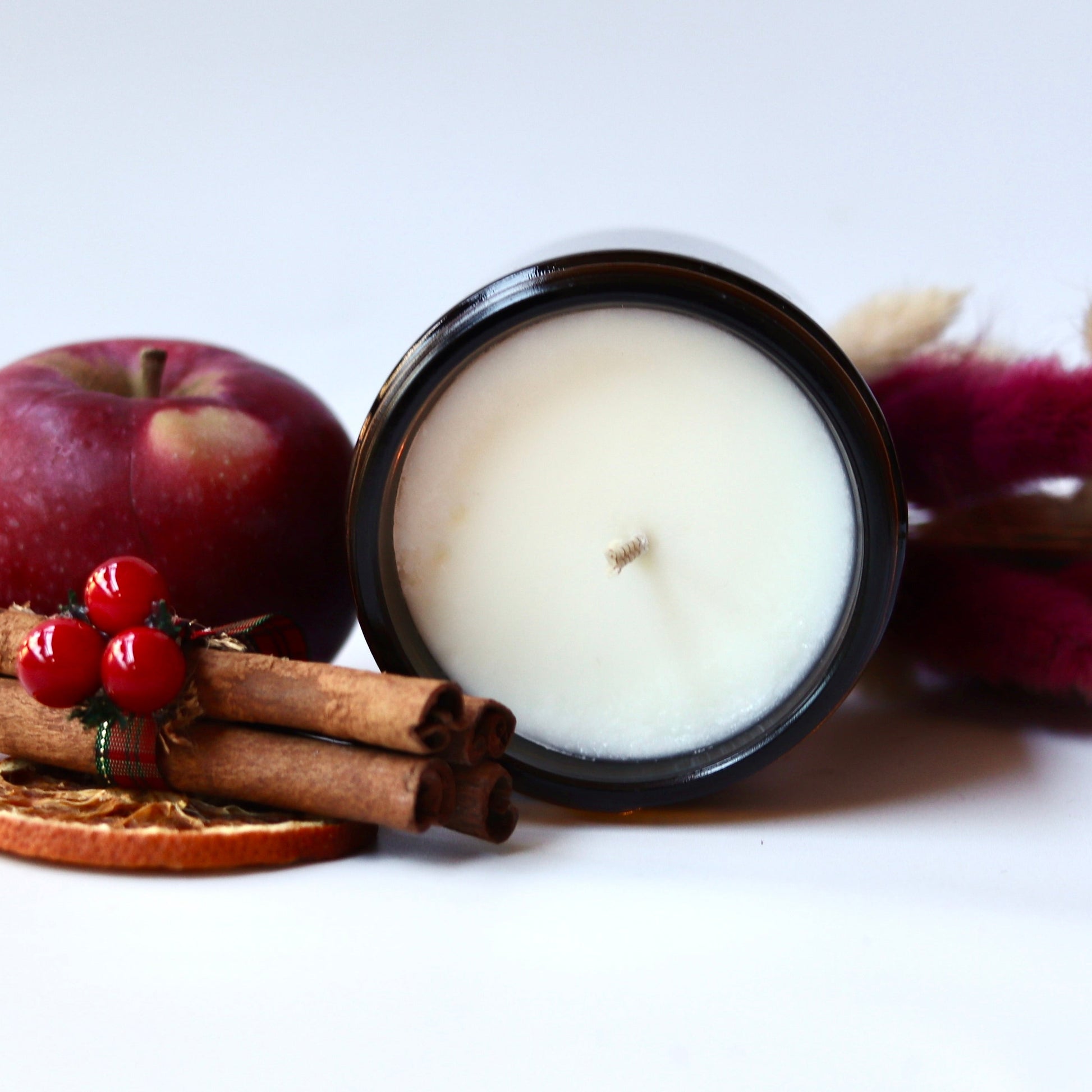 Candle in a black container with an apple, cinnamon sticks, and dried flowers on a white background