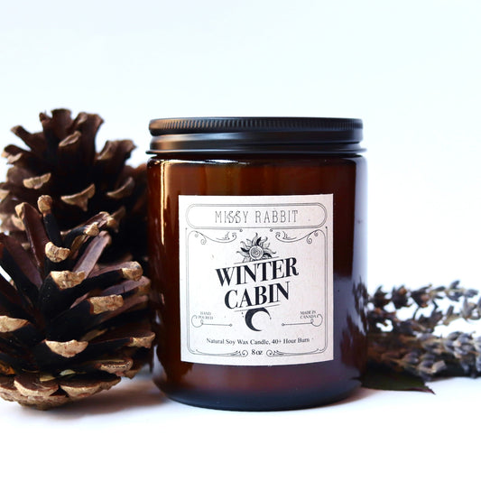 Candle labeled 'Winter Cabin' with a pine cone and lavender on a white background