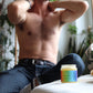 A shirtless man sitting on a white lounge chair in a plant-filled room, with a small jar of body butter in the foreground on a white surface.